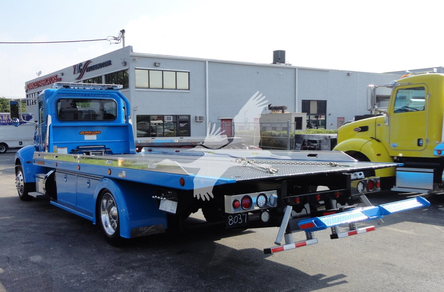 Peterbilt 337 rollback truck in blue with flatbed rear and hydraulic ramps, shown from side/rear view in a dealership lot.