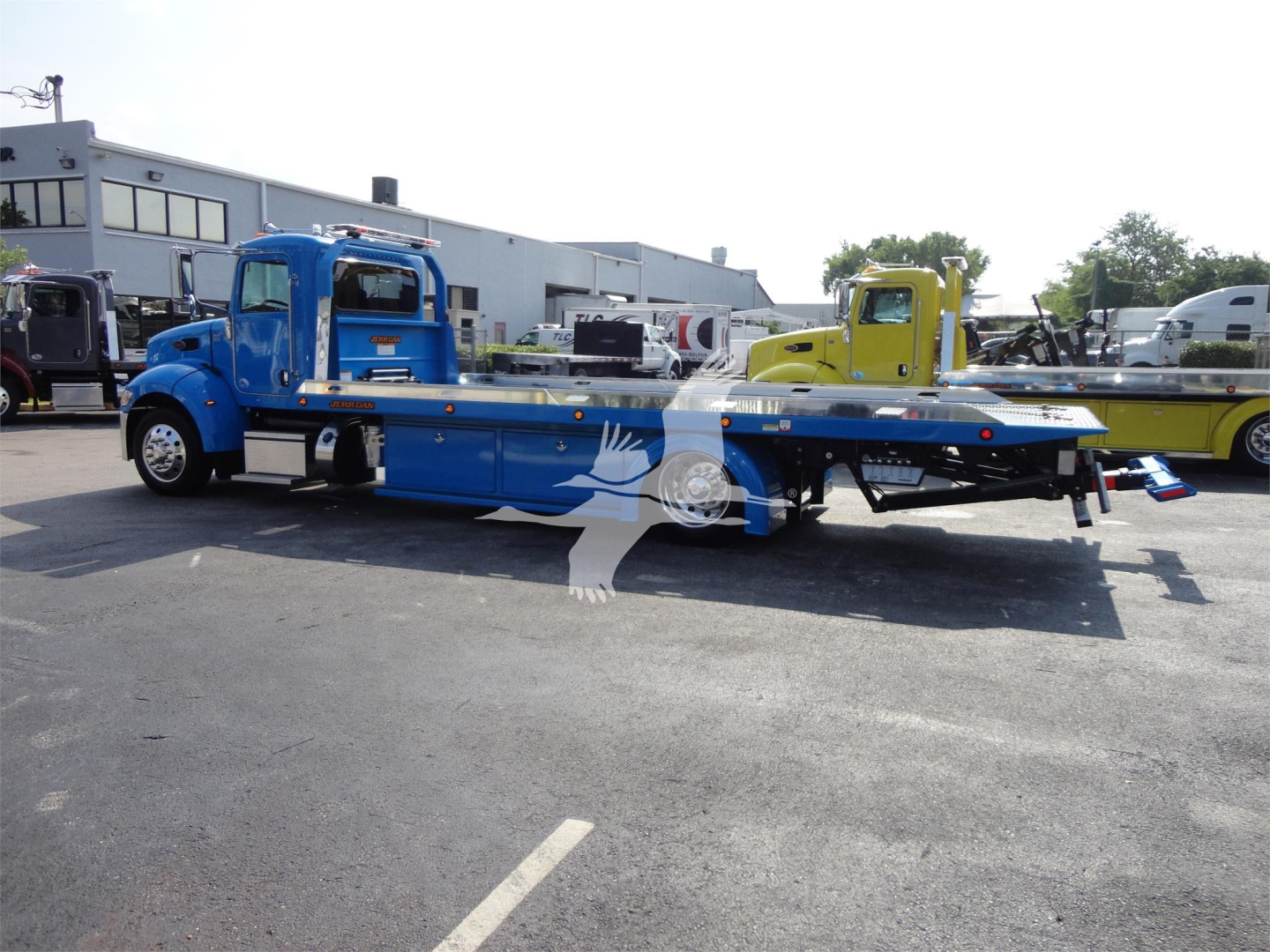 Blue Peterbilt 337 rollback tow truck with flatbed, side view showing cab and long bed, yellow and other trucks in background.