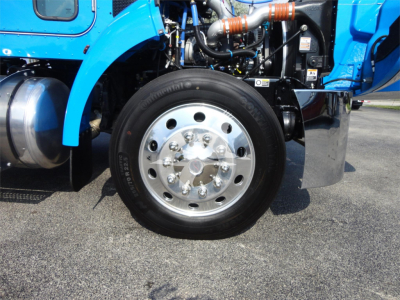 Front wheel of a blue 2022 Peterbilt 337 rollback truck with polished aluminum rim and visible engine bay components.