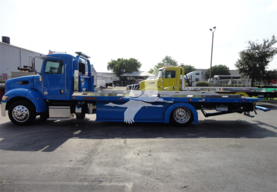 Peterbilt 337 rollback truck in blue and white with flatbed, side view showing the vehicle and a second yellow truck in background.