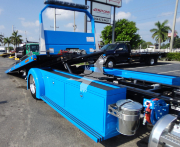 Blue Peterbilt 337 rollback truck body with hydraulic wheel lift and tool storage compartments. Side view showing blue cabinetry, metal rollers, and a silver canister near the chassis.