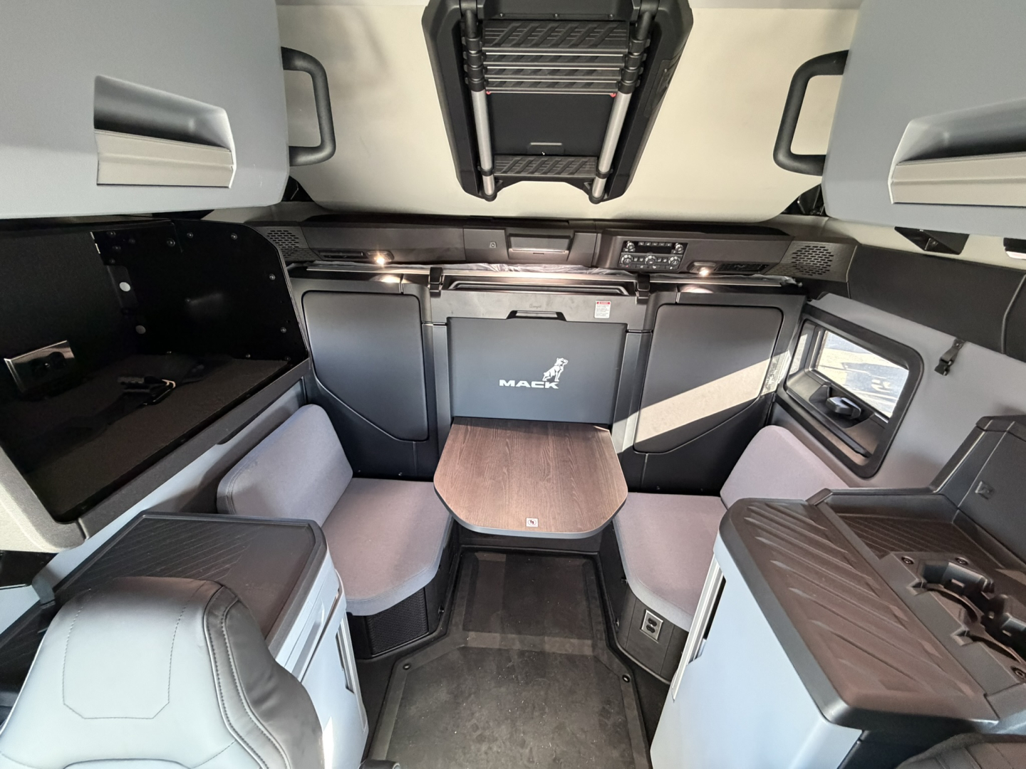 Cabin interior of a 2026 Mack PR64T76H showing bunk-style seating, table, and overhead storage with Mack logo on panels.