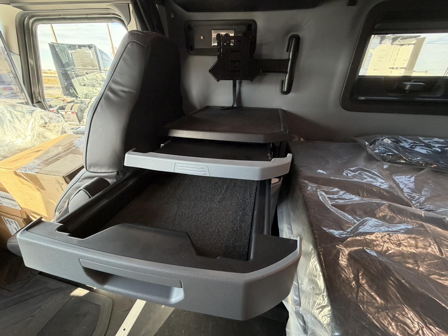 Truck cab interior showing a gray two-tier storage drawer system; side window, bed with plastic covering, and a mount/seat area visible in a Mack PR64T76H truck.