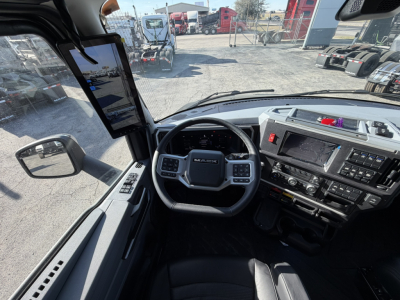 Cab view of a Mack PR64T76H truck showing steering wheel, dashboard with touchscreen, and side mirror.