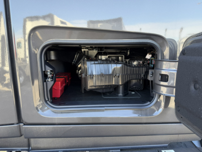 Image of a Mack truck PR64T76H engine bay visible through an open side door panel, with a chrome engine cover and red battery visible on left.