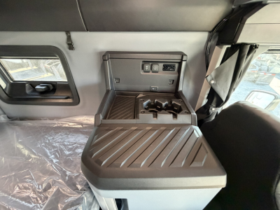 Mack PR64T76H truck interior center console with cup holders and storage, control panel, and window visible; cabin area showing overhead shelf and side window.