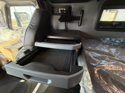 Truck cab interior showing a gray two-tier storage drawer system; side window, bed with plastic covering, and a mount/seat area visible in a Mack PR64T76H truck.