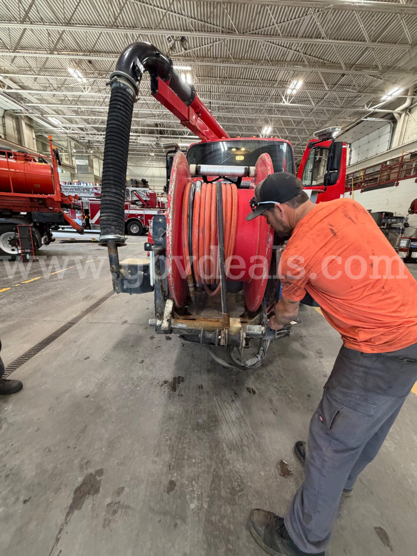 Industrial workshop with a Freightliner 114SD and a large red hose reel on a truck. Technician in orange shirt and cap bends near the hose reel, others and equipment visible in background.