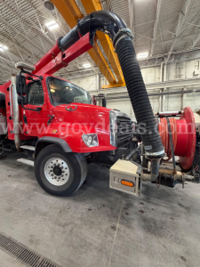 Freightliner 114SD vacuum truck front-left view with large black flexible hose and rear vacuum unit; red cab and chassis, overhead crane in workshop.
