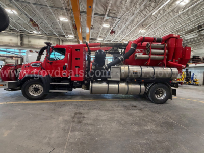 Freightliner 114SD vacuum truck in a workshop showing the side profile with a red cab and vacuum tank, hoses, and metallic components. Visible text includes ‘VAC’ on the equipment hose assembly and engine bay labels near the cab.