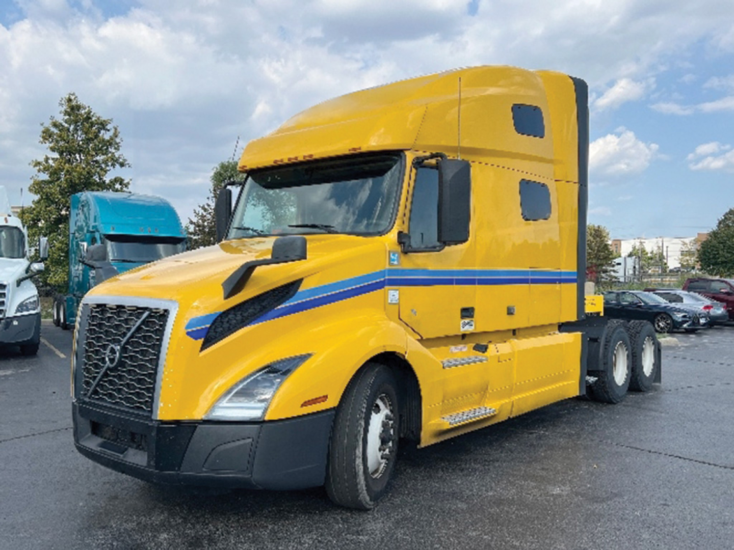 Truck engine bay and cab of a yellow 2022 Volvo VNL 760 conventional sleeper, side view showing Volvo grille, blue stripe, and cab details.