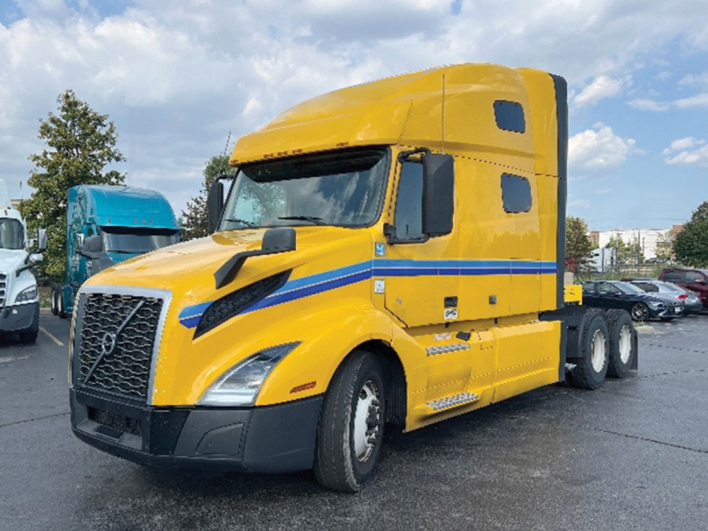Truck engine bay and cab of a yellow 2022 Volvo VNL 760 conventional sleeper, side view showing Volvo grille, blue stripe, and cab details.