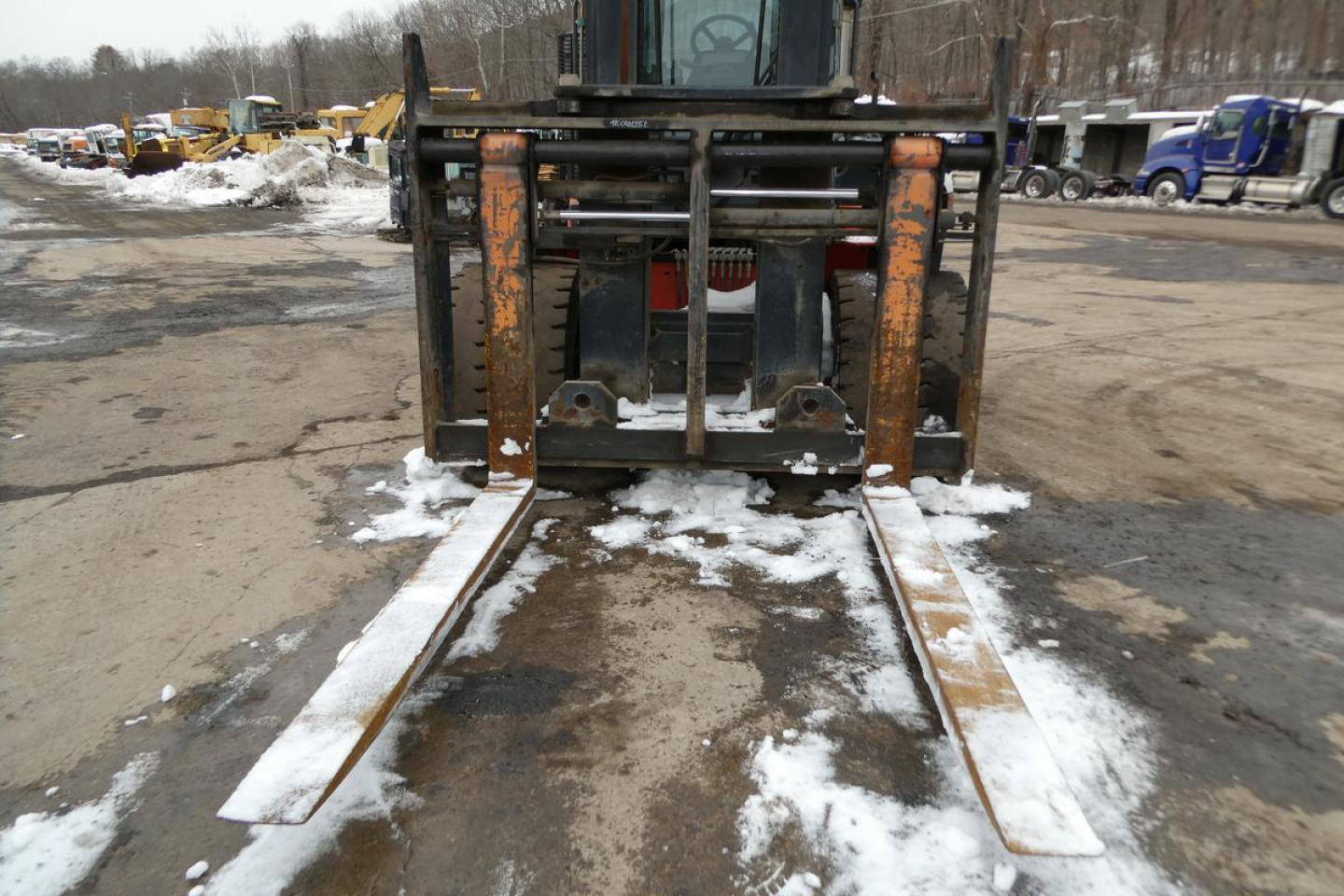 Forklift forks extended forward with rusted carriage in front, snow on ground, industrial yard in background.