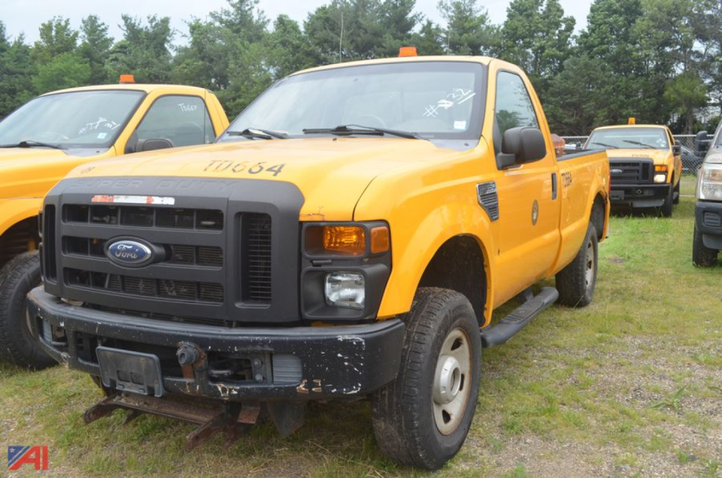 Ford F-250 Super Duty dump truck in yellow, front view, with black grille and bumper, worn front hitch, in a lot with other trucks