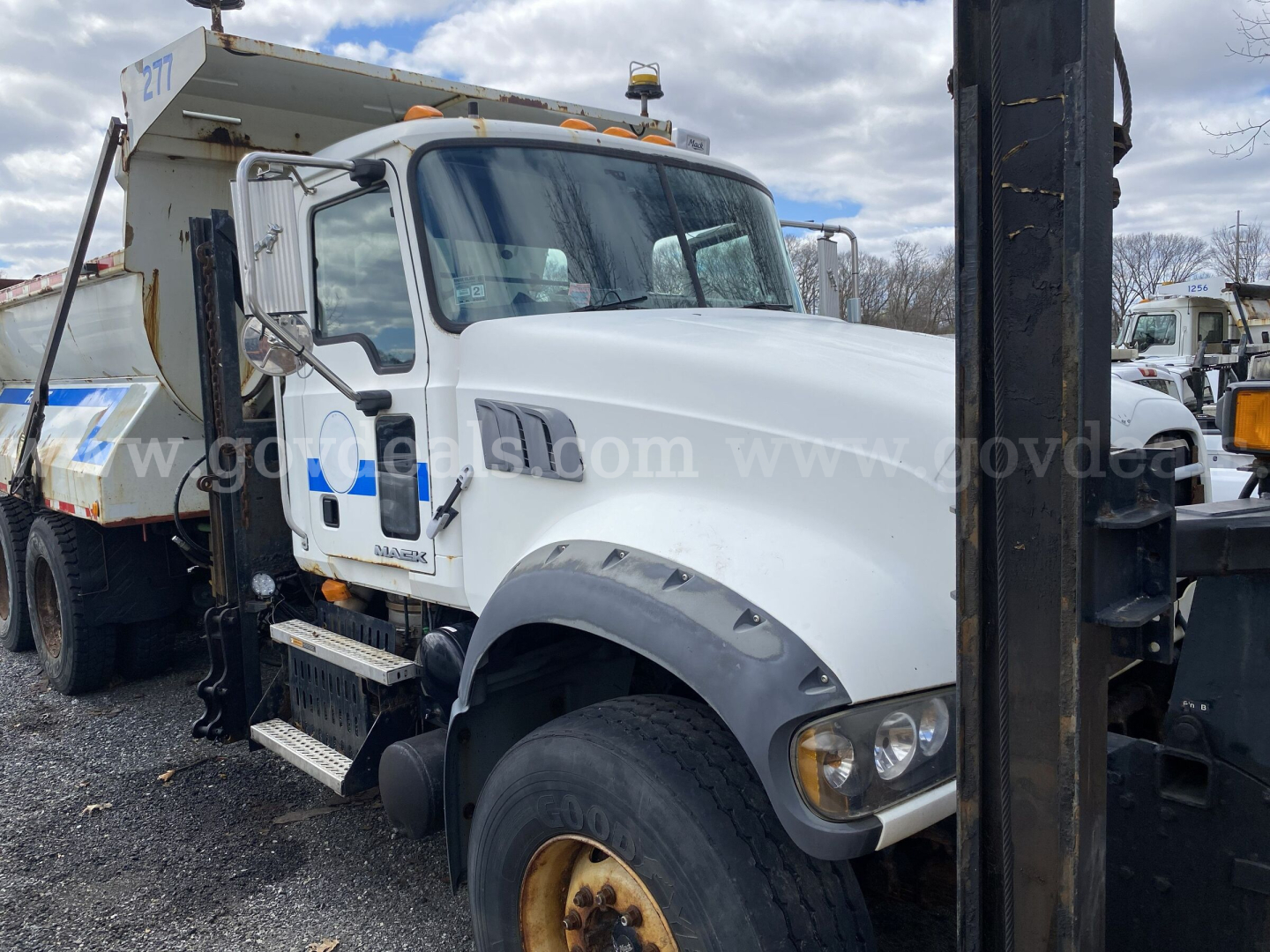 Mack GU713 dump truck with white cab, front view showing side steps and large fender, VIN plate not visible, heavy equipment in background.