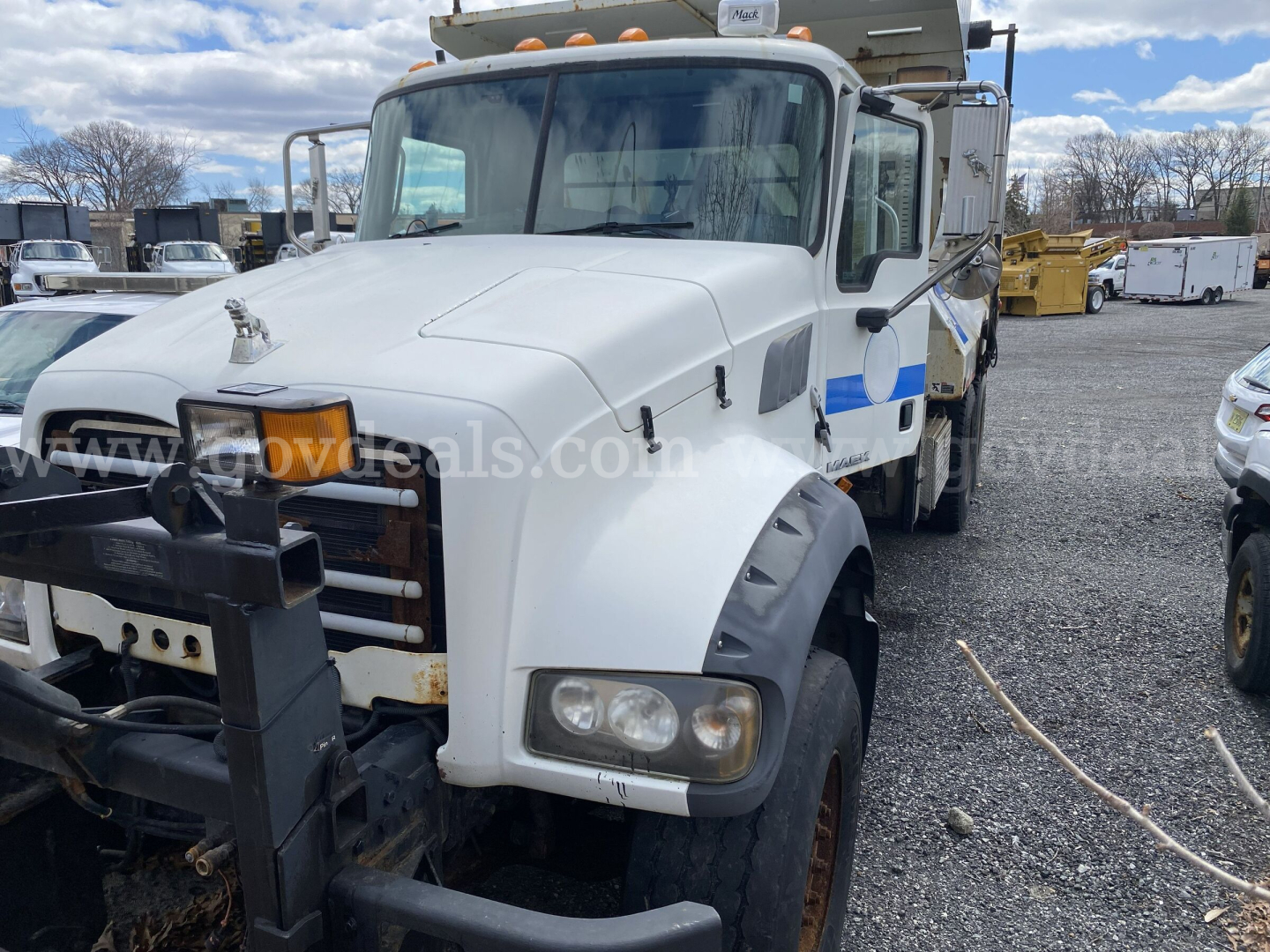 Mack GU713 dump truck front view showing white cab and dual headlights, with plow attachment and warning lights on top.