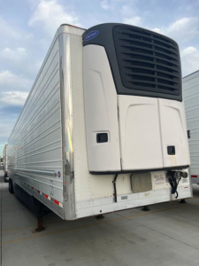 Trailer rear view showing refrigeration unit on top of a white refrigerated trailer with black lower vent grille and rear doors closed.