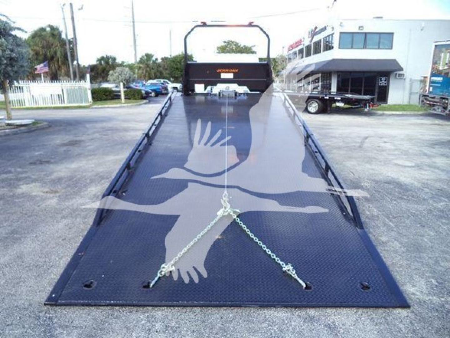 Truck rollback bed with Black steel platform and chains in foreground, Hino branding on cab, dealership building in background.