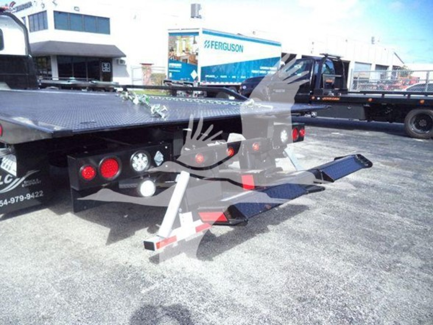 Rear view of a 2025 Hino 268A rollback tow truck with hydraulic flatbed and ramps, parked in a lot with other trucks in the background.