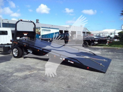 2025 Hino 268A rollback truck on a lot with a long folded flatbed trailer and black frame; background shows dealership buildings and other vehicles.