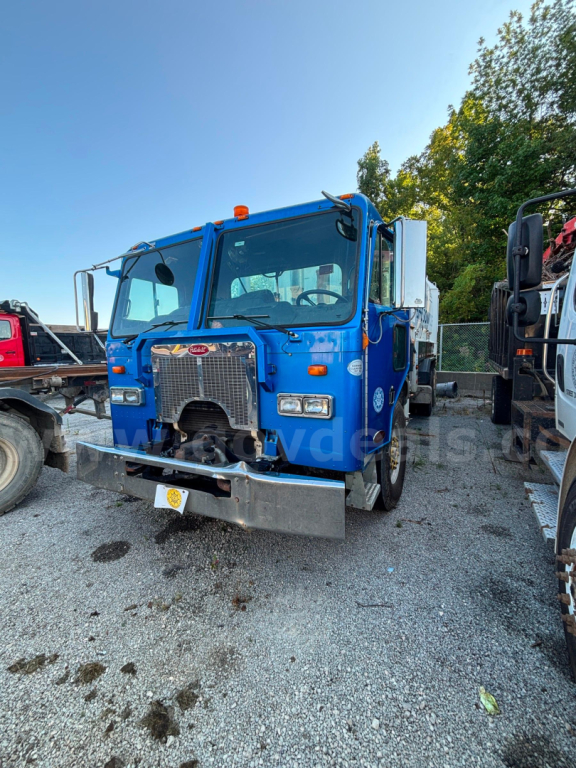 Blue Peterbilt COE truck front view showing chrome grille, bumper, and dual headlights; VIN sticker not visible; gravel lot with other trucks in background.