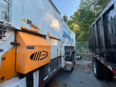 Truck body side with orange hydraulic unit and 'JEFFERSON' branding, side rail, and rust spots visible on a garbage truck in a lot.