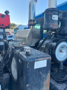 Truck engine bay with hydraulic system; label 'HYDRAULIC' on metal container; visible gears, hoses, and a pressure gauge area; rust patches; various stamped stickers.