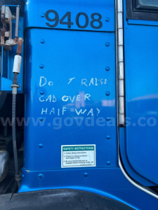 Blue Peterbilt truck cab side with large black number 9408, handwritten white text reading Do not raise cab over half way, safety instructions sticker at bottom, riveted panel and hoses.