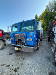 Blue Peterbilt COE truck front view showing chrome grille, bumper, and dual headlights; VIN sticker not visible; gravel lot with other trucks in background.