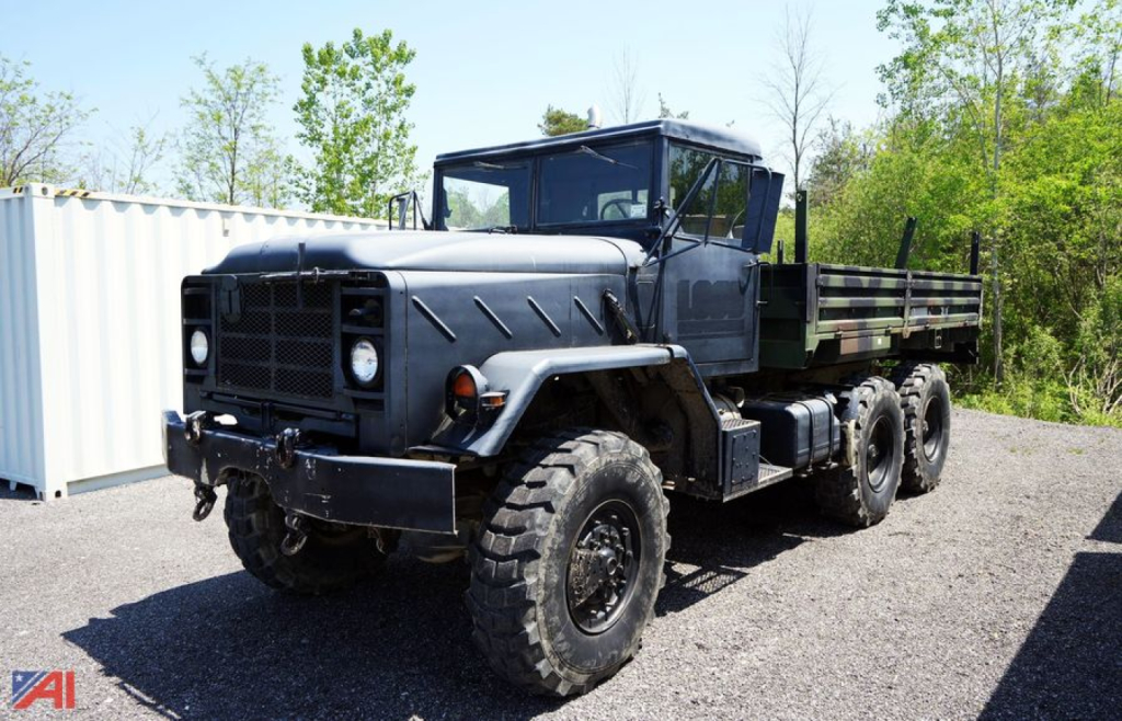 Military 1990 BMY 14 truck in a yard with six wheels and flatbed, matte gray paint and rugged off-road tires.
