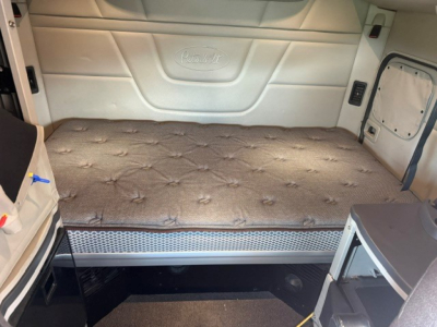 Peterbilt 579 sleeper cab interior with a raised bed mattress and side storage; light gray panels and embossed Peterbilt logo on rear wall.
