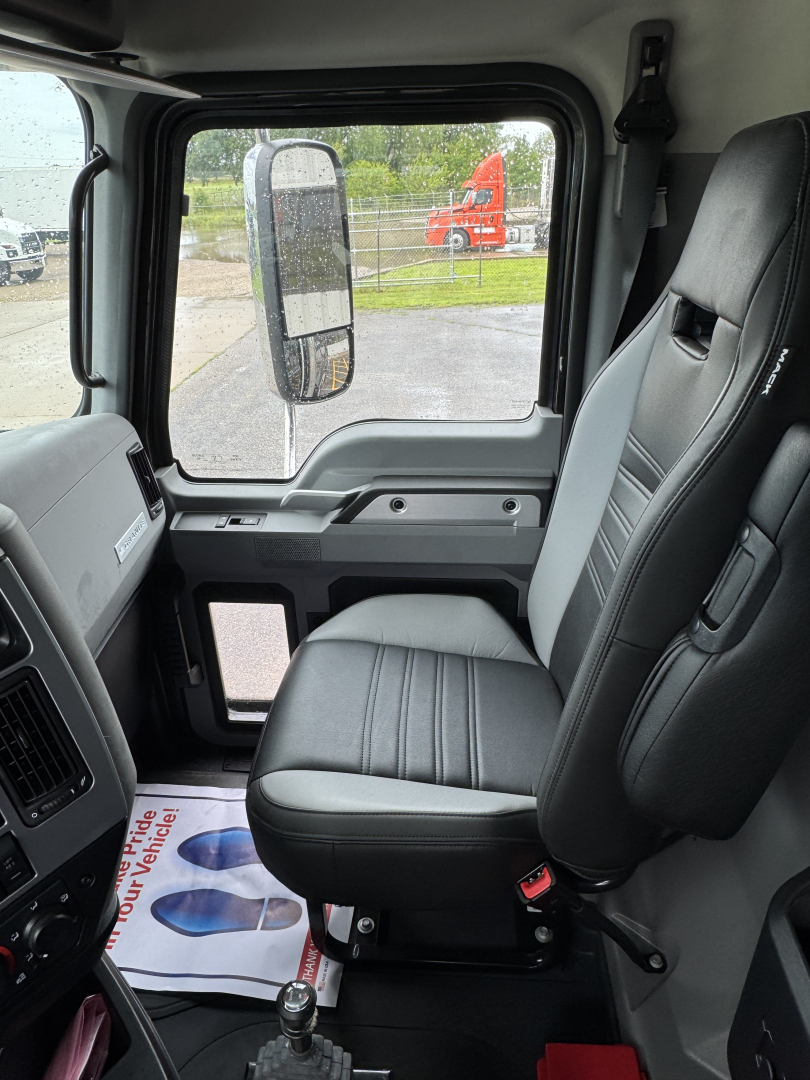 Interior cab view of a 2025 Mack GR104F dump truck showing the driver seat, side mirror, and door panel with control area; dashboard and windshield visible with exterior truck in background.