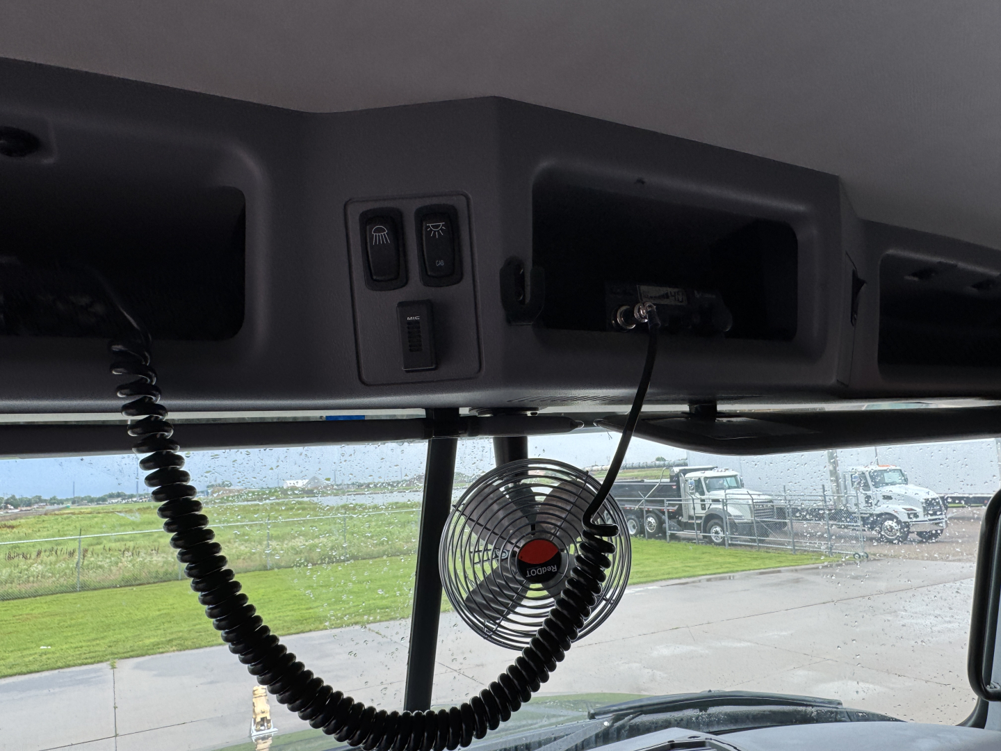 Cab interior overhead area with two control switches, a mounted microphone or handheld mic, and a small circular fan. Window view shows a parking lot and trucks in background.