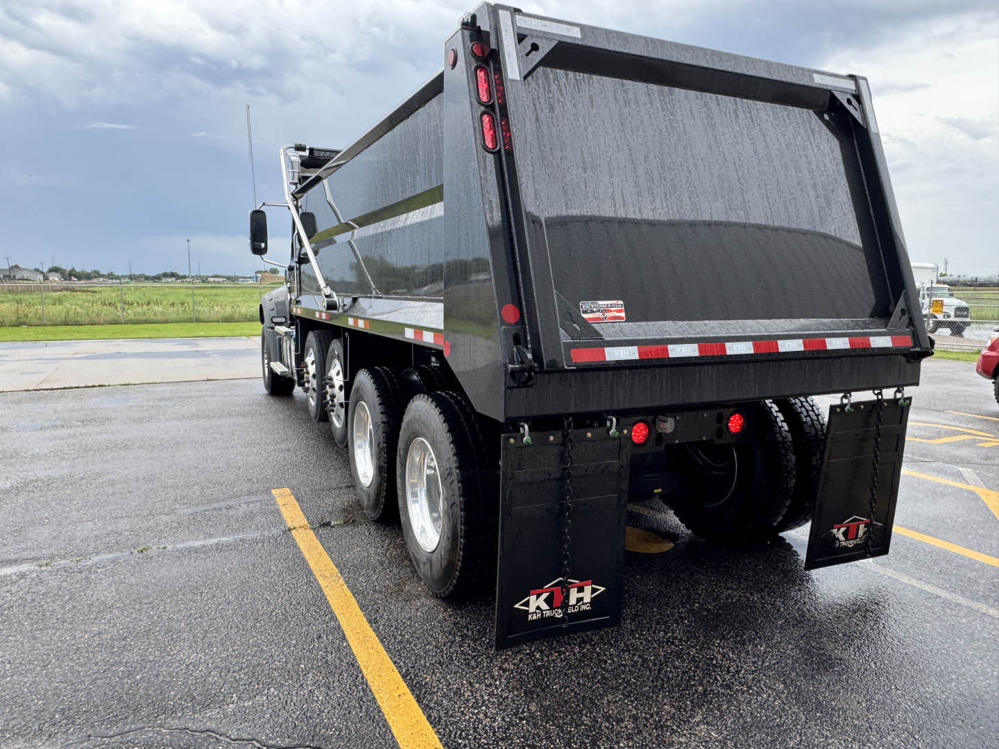 Dump truck rear view showing black dump bed with reflective tape, triple rear axles, mud flaps, and tailgate. Visible branding: 4KH mud flaps.