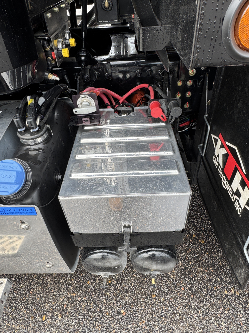 Truck battery compartment with two large cylindrical terminals, red and black cables, and a rectangular metal battery box mounted under a Mack GR104F truck.