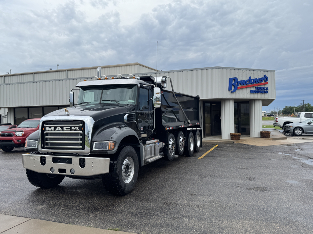 Truck image: 2025 Mack GR104F dump truck in dark color, front grille with MACK letters, three-axle rear, parked in lot near dealership building.