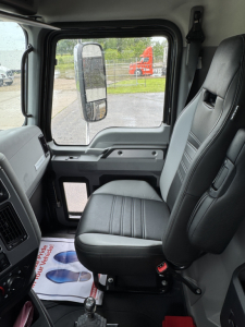 Interior cab view of a 2025 Mack GR104F dump truck showing the driver seat, side mirror, and door panel with control area; dashboard and windshield visible with exterior truck in background.