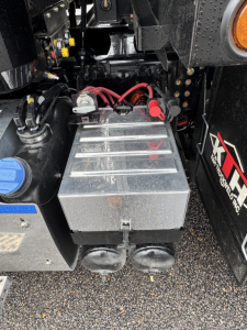 Truck battery compartment with two large cylindrical terminals, red and black cables, and a rectangular metal battery box mounted under a Mack GR104F truck.