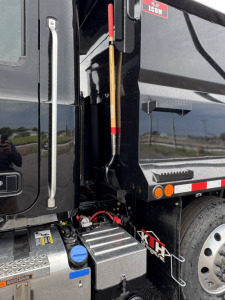 Truck cab and dump body area showing step deck, hydraulic lever, and blue coolant fill cap near the chassis.