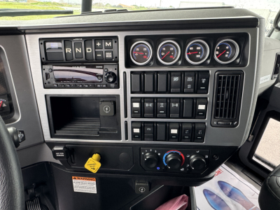 Dashboard of a 2025 Mack GR104F dump truck showing instrument gauges, control switches, climate controls, and radio. Clear text on knobs and buttons visible.