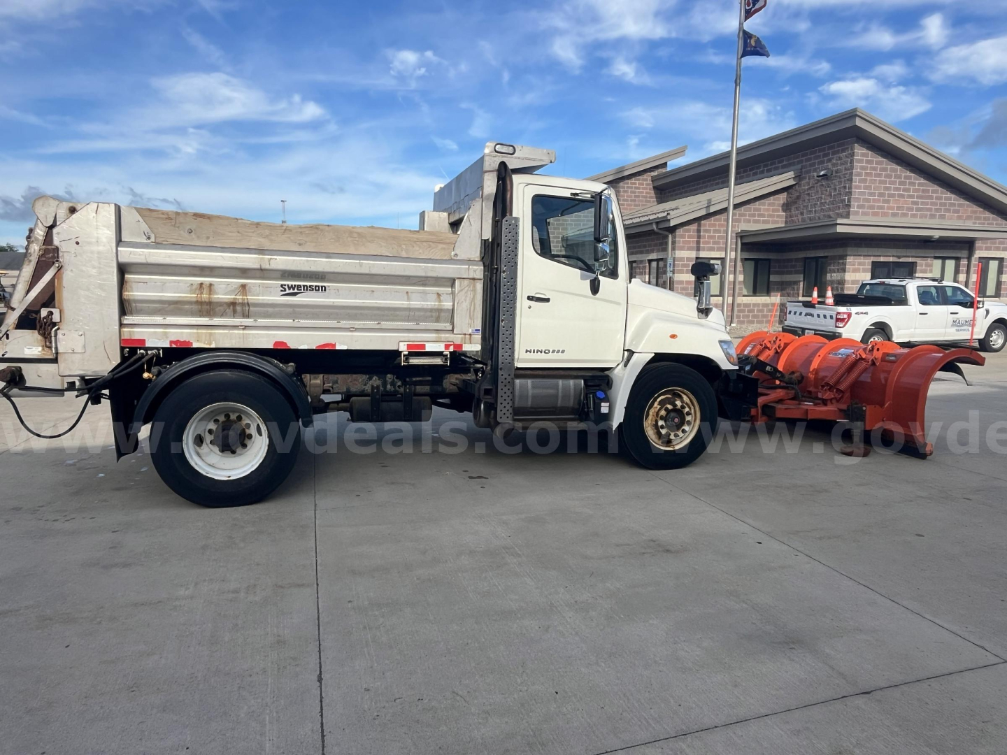 White 2012 Hino 338 dump truck with snowplow attachment in foreground, side view showing dump bed and wheel, dealership lot in background.