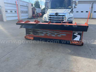 Truck front view with snow plow attachment in foreground and Hino cab in background; plow edge and orange stakes visible.