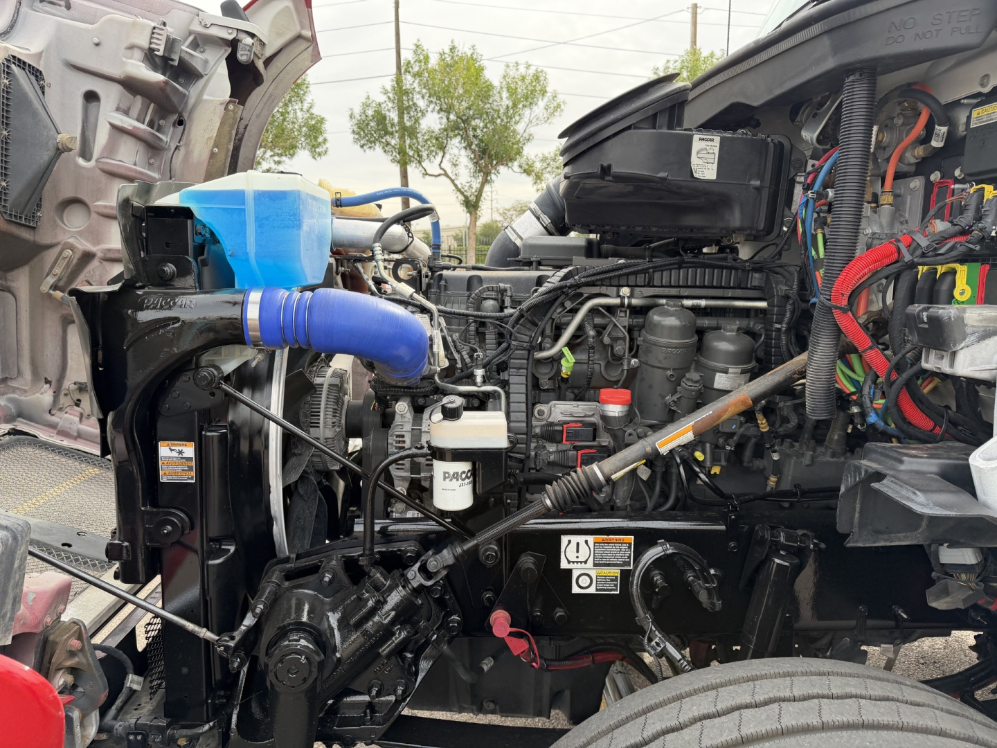 Kenworth T880 engine bay with exposed components including blue coolant reservoir, hoses, wiring, and hydraulic lines; visible VIN plate area and label near center.