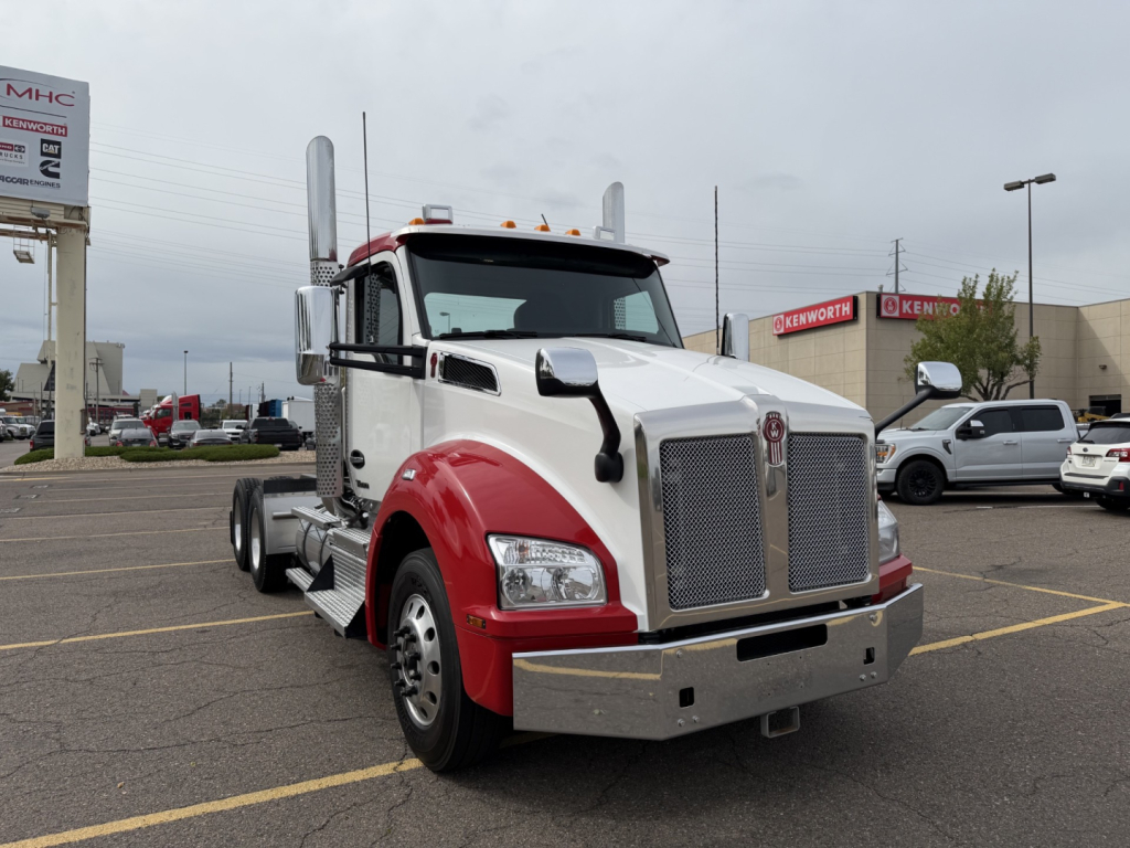 Kenworth T880 conventional daycab truck in a parking lot, front three-quarter view, white cab with red fenders, large chrome grille, dual tall exhaust stacks, chrome bumper.