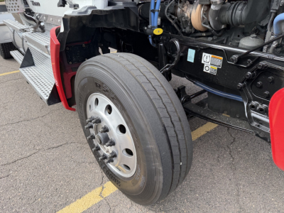 Kenworth T880 front-left wheel and engine bay view showing steering linkage, exposed engine components, and warning labels.