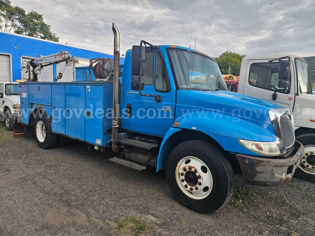 Blue 2004 International 4300 service/utility truck shown in side and front view with equipment on the flatbed and crane. Clear cab, large mirrors, exhaust stack visible on passenger side, front bumper and wheel.
