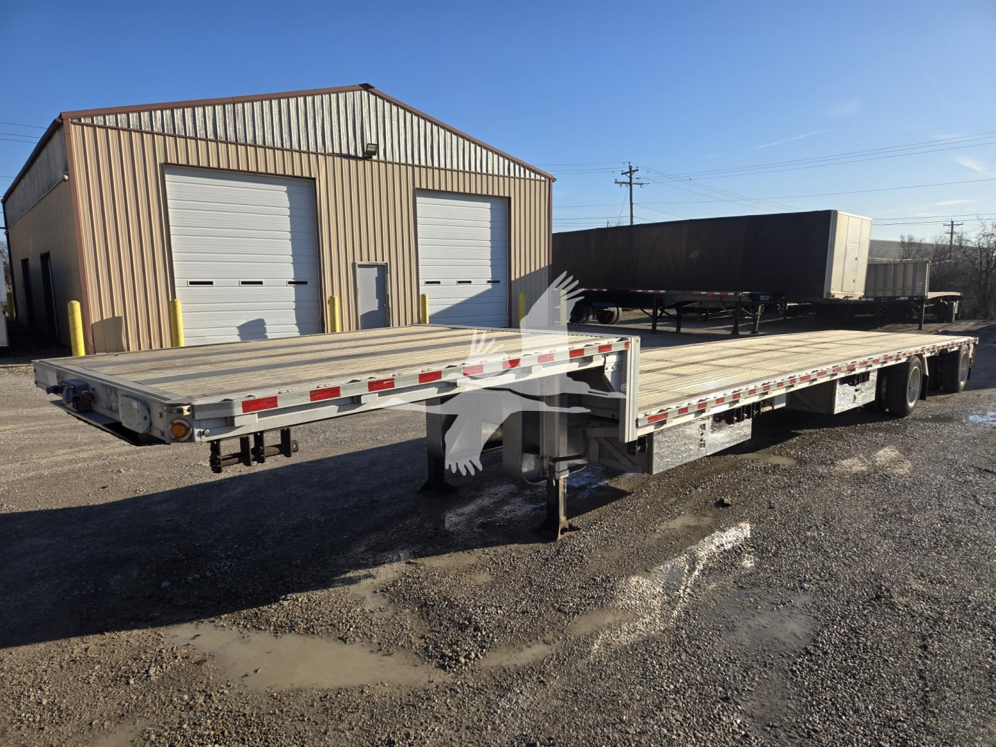Flatbed trailer in front of a metal shop showing drop deck trailer and dual rear axles with reflective tape.