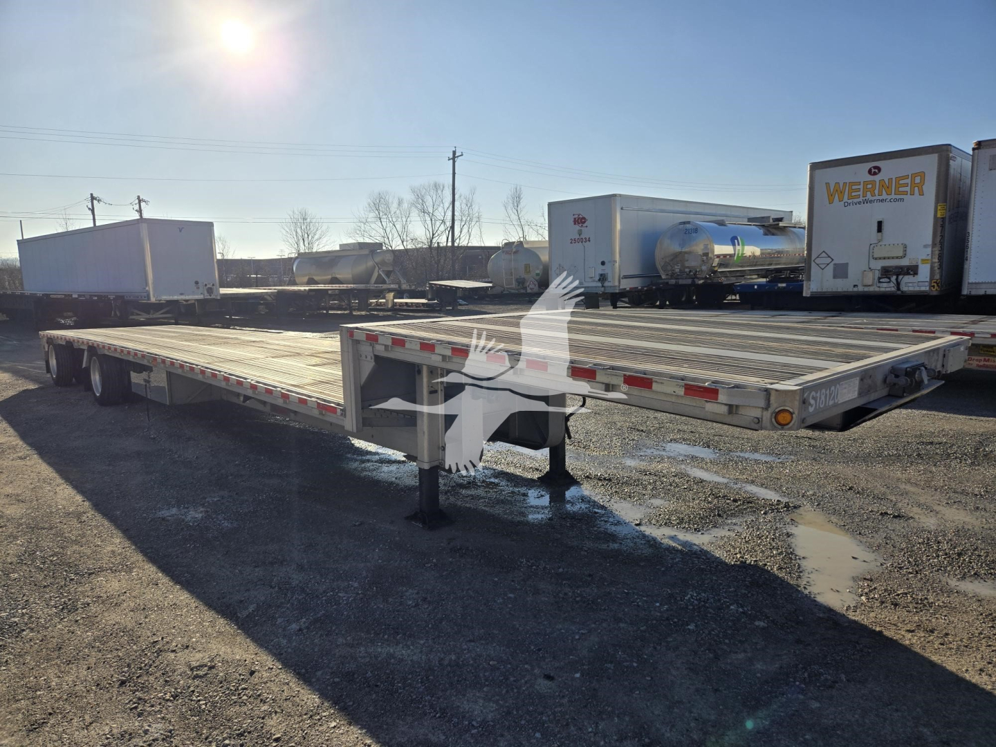 Drop deck trailer (flatbed) with extended gooseneck area, low ground clearance, and landing legs visible. Side rails and reflective tape along edges; background shows other trailers.