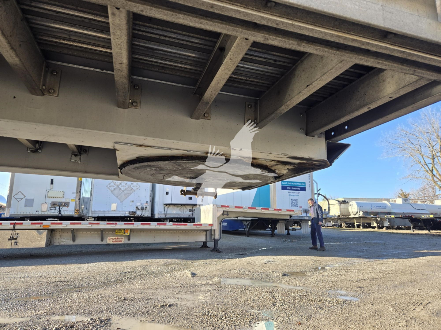 Trailer underside showing drop deck mechanism of a 2019 Reitnouer Dropmiser trailer with supporting beams and ground crew in background.