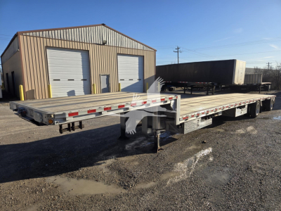 Flatbed trailer in front of a metal shop showing drop deck trailer and dual rear axles with reflective tape.